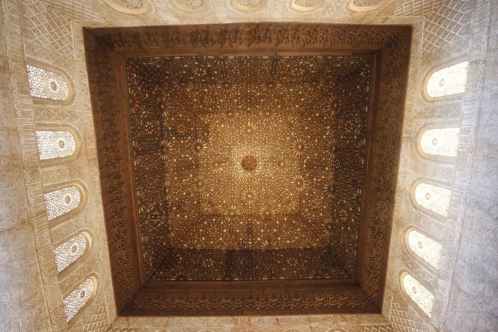 A square carved wooden ceiling with star patterns, surrounded by pierced stucco windows, in the Hall of the Ambassadors at the Alhambra in Granada