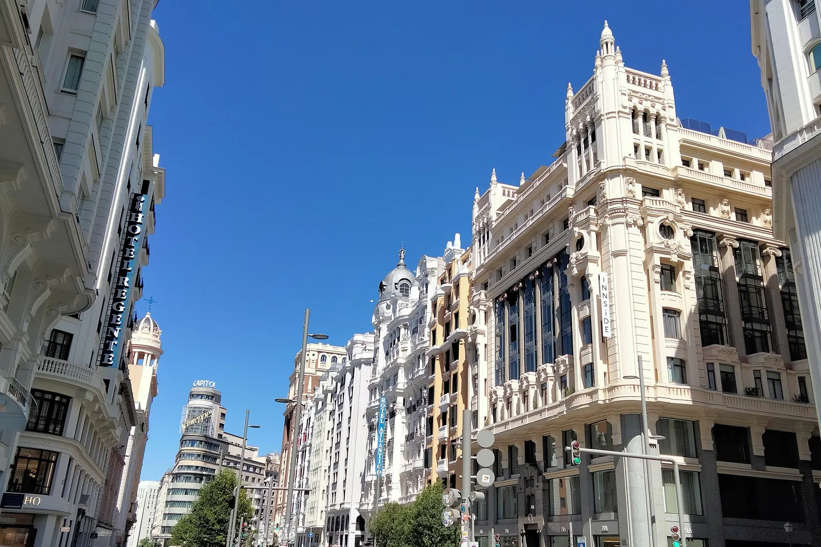 White and cream buildings line a sunlit stretch of Gran Vía in Madrid under a deep blue sky, with the Capitol building in the distance